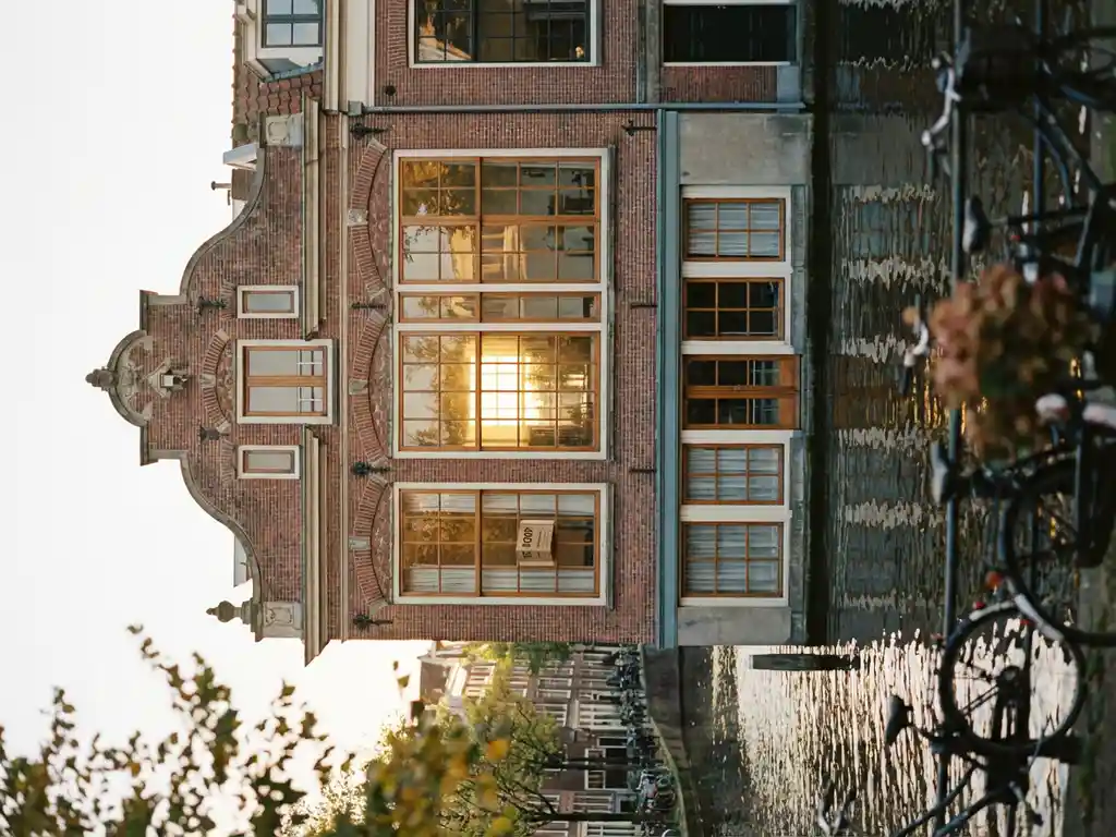 Amsterdams grachtenpand met terracottakleurige bakstenen gevel, grote schuiframen en een "Te Koop"-bord in warm middaglicht.