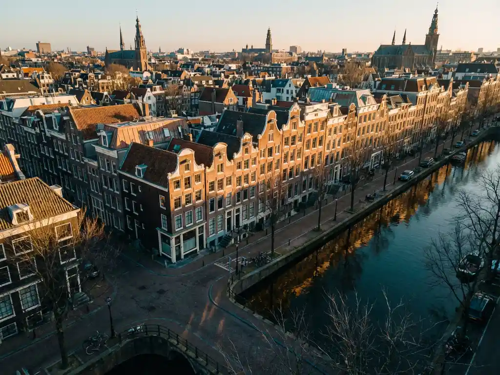 Luchtfoto van Amsterdamse grachtenpanden met terracotta daken en gouden gevels in warm ochtendlicht, weerspiegeld in het stille kanaalwater.