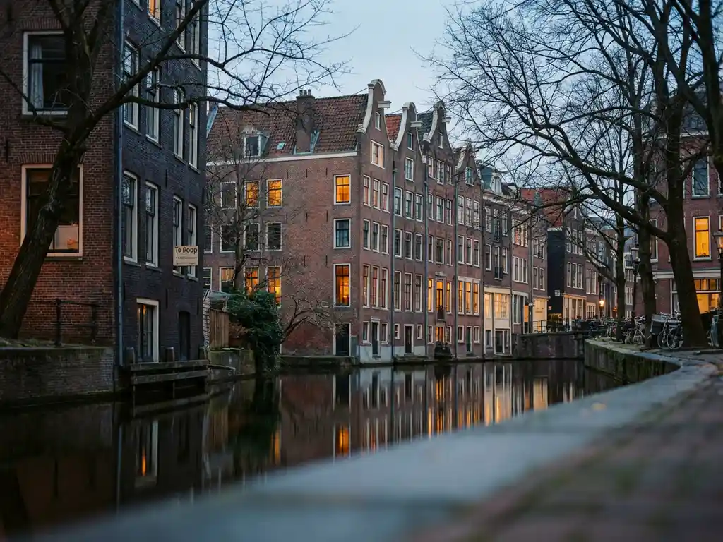 Amsterdamse grachtenpanden met terracottarode gevels weerspiegeld in donker water, kale winterbomen en een Te Koop-bord bij schemerlicht.