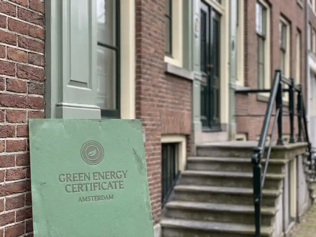 Groen energielabel certificaat tegen de bakstenen gevel van een Amsterdams grachtenpand met stenen trap en ijzeren leuning.